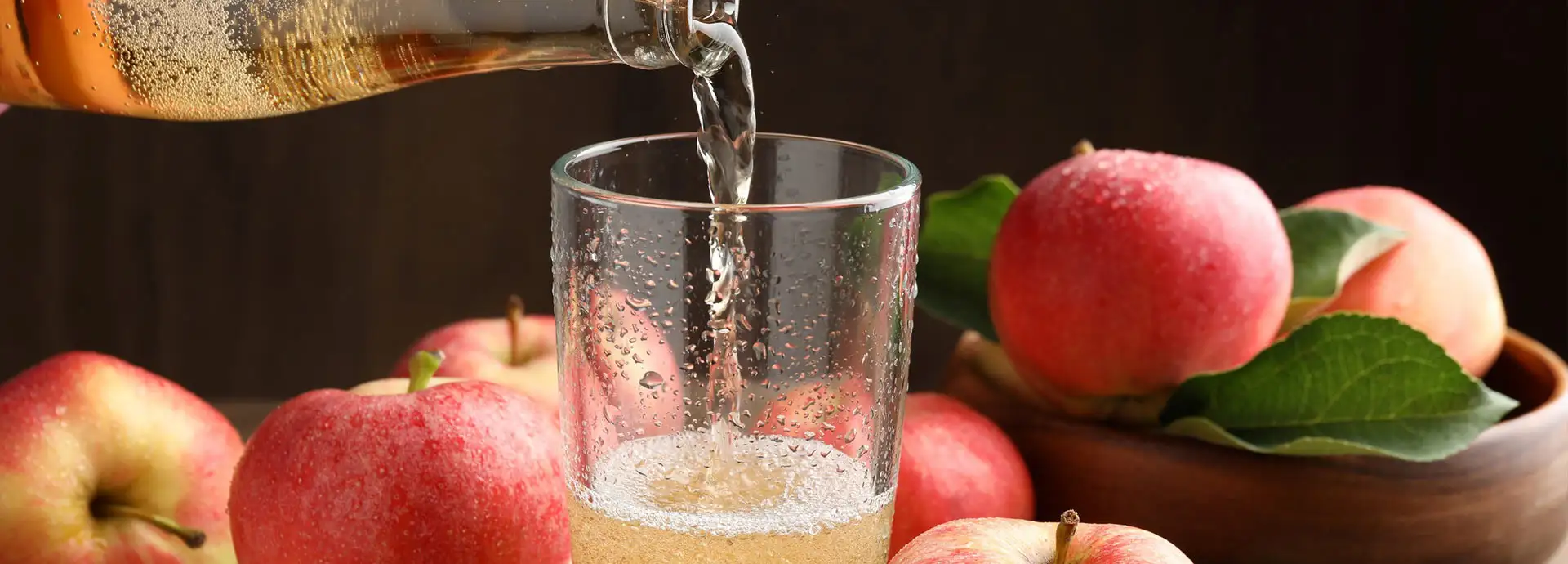 Image of a bottle of an alcoholic liquid pouring into a glass that is surrounded by red apples.